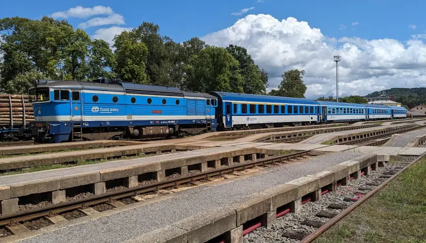 Blauer cz train mit mehreren Waggons steht auf Gleisen, umgeben von Bäumen und einem blauen Himmel mit Wolken.