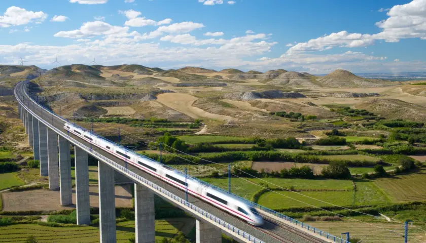 Schneller Zug auf einer Brücke in Spanien, der durch eine hügelige Landschaft mit Windrädern fährt.