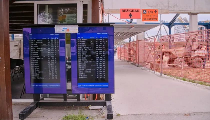 Anzeigetafeln am Bahnhof Ljubljana zeigen Abfahrts- und Ankunftszeiten. Baustellenabsperrungen und ein Bagger sind im Hintergrund sichtbar.