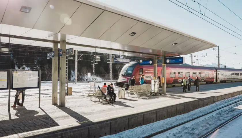 Winterliche Szene am **Lech Bahnhof**. Ein roter Zug steht bereit, während Reisende auf dem verschneiten Bahnsteig warten.