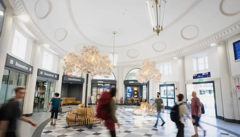 Menschen eilen durch den hellen, modernen **Coburger Bahnhof** mit seinen auffälligen Lampen und Sitzgelegenheiten.