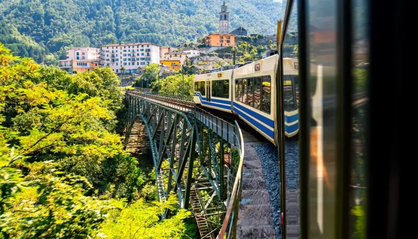 Mit dem Zug in die Toskana: Eine malerische Fahrt über eine Eisenbahnbrücke, vorbei an grünen Hügeln und einem charmanten Dorf mit Kirchturm.