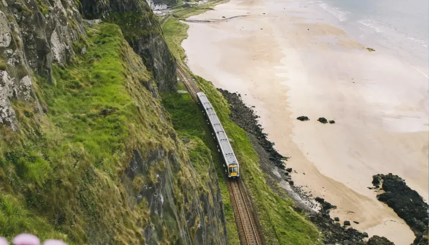 Ein Zug fährt entlang der Küste von **Railway Ireland**. Grüne Hänge stürzen zum Strand ab, das Meer glitzert in der Ferne.