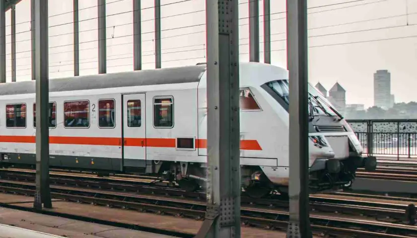 Moderner weißer IC-Zug mit orangefarbenen Streifen fährt auf Gleisen über eine Brücke. Im Hintergrund ist eine Stadt zu sehen.