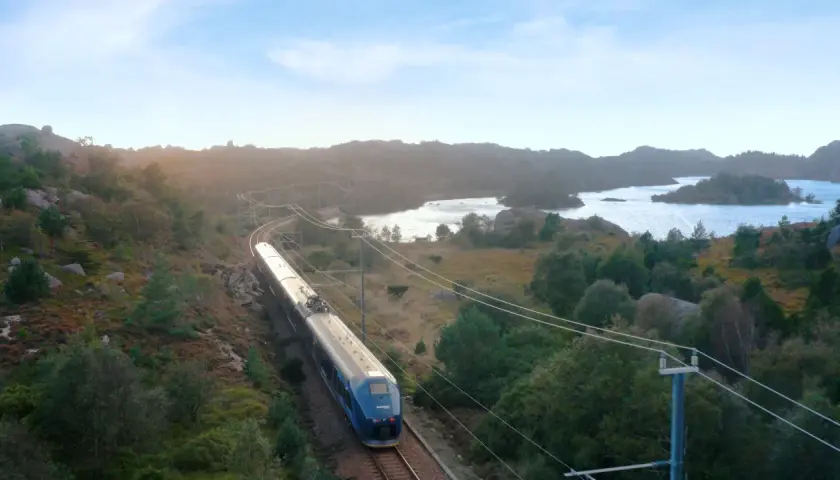 Ein blauer Zug fährt auf der Strecke Oslo-Stavanger durch eine hügelige, grüne Landschaft mit Seen und Felsen.