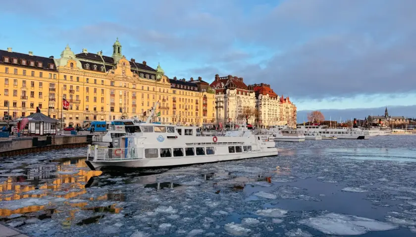 Eisbedeckter Kanal in Stockholm mit einem weißen Schiff, das an einer Promenade mit historischen Gebäuden vorbeifährt. Ein blauer Bus, der an einen schwedischen Zug erinnert, ist im Hintergrund zu sehen.