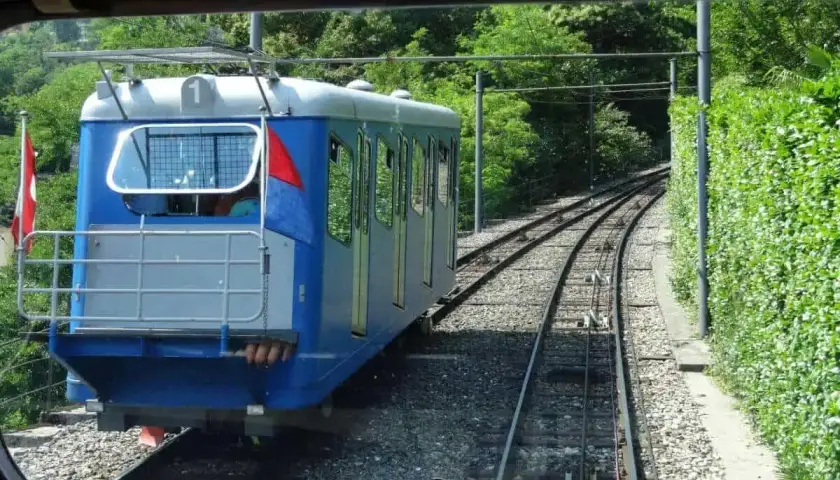 Mit dem Zug zum Lago Maggiore: Eine blaue Standseilbahn fährt auf Schienen durch üppiges Grün.