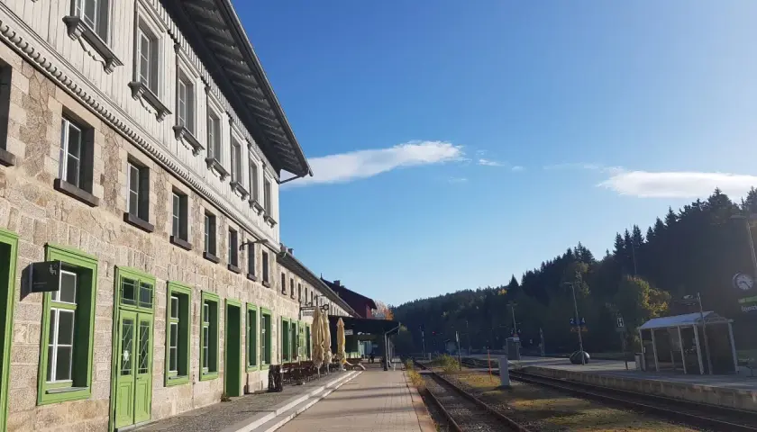 Der **Bayerisch Eisenstein Bahnhof** mit seinem historischen Gebäude und den grünen Fensterrahmen, umgeben von einer Waldkulisse unter blauem Himmel.