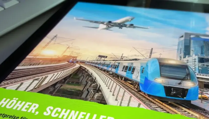 Blauer Hochgeschwindigkeitszug fährt auf erhöhter Strecke, während ein Flugzeug am Himmel schwebt. Eine Anzeige wirbt für "Zug statt Flug".