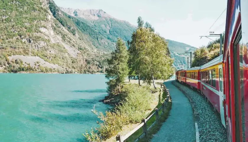 Ein roter und gelber Fernzug fährt entlang eines türkisfarbenen Sees und majestätischer Berge.