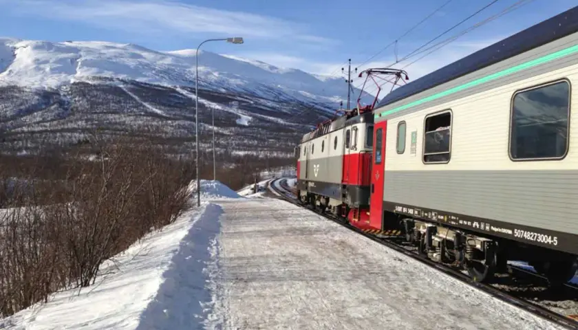 Der Zug von Stockholm nach Kiruna fährt durch eine verschneite Winterlandschaft mit schneebedeckten Bergen im Hintergrund.