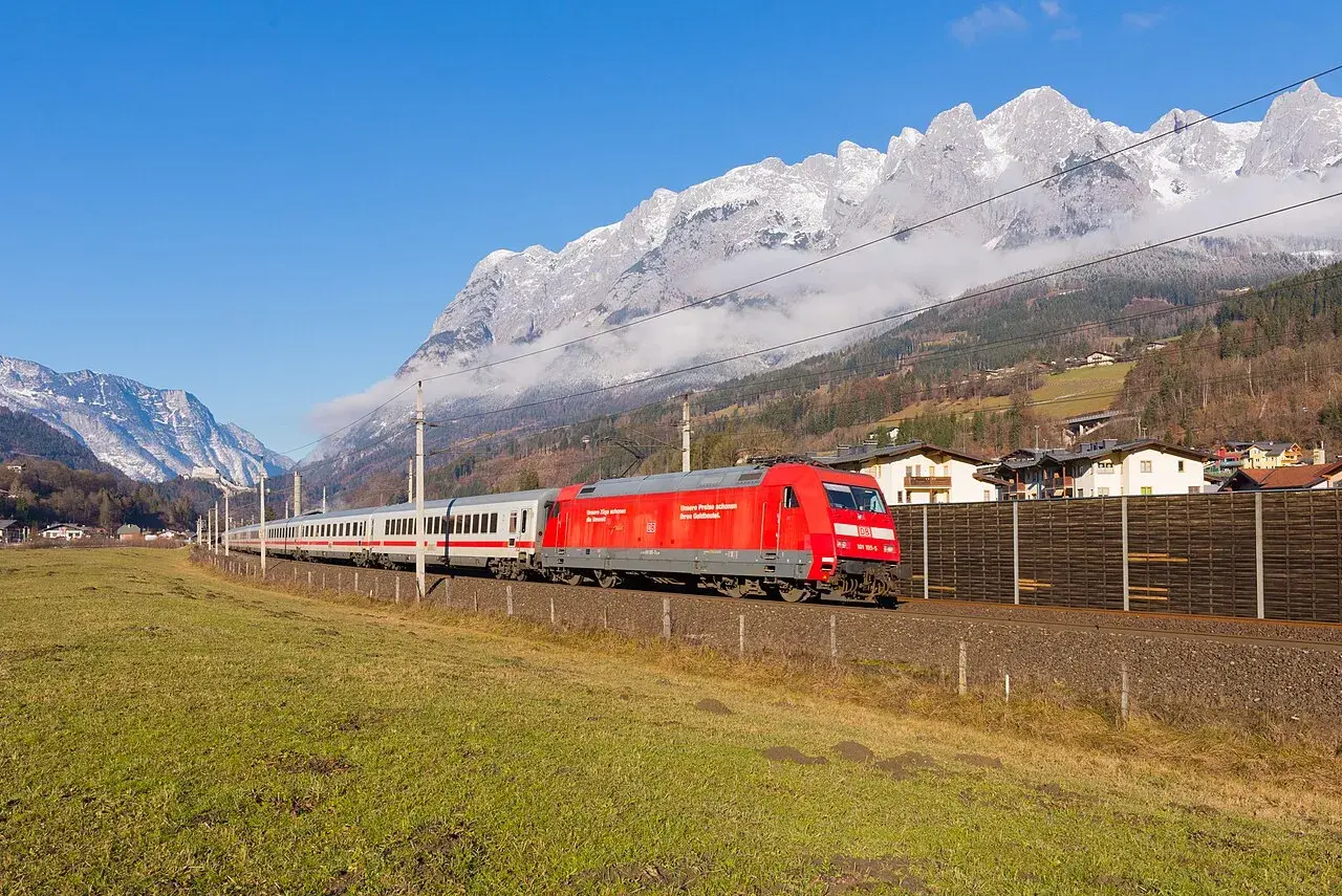 Ein roter Eurocity-Zug fährt durch eine grüne Wiese, im Hintergrund schneebedeckte Berge und ein Dorf.