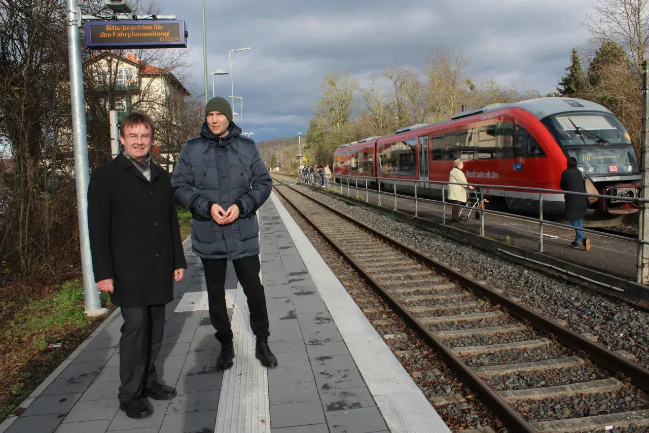 Zwei Männer stehen auf dem Bahnsteig des kleinsten Bahnhofs Deutschlands. Ein roter Zug der Westfrankenbahn wartet auf Fahrgäste.