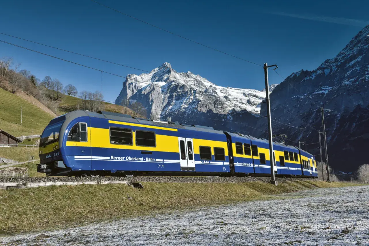 Blauer und gelber Zug der Berner Oberland-Bahn fährt durch eine verschneite Landschaft, mit schneebedeckten Bergen im Hintergrund. Die Fahrt von Interlaken nach Lauterbrunnen beginnt.