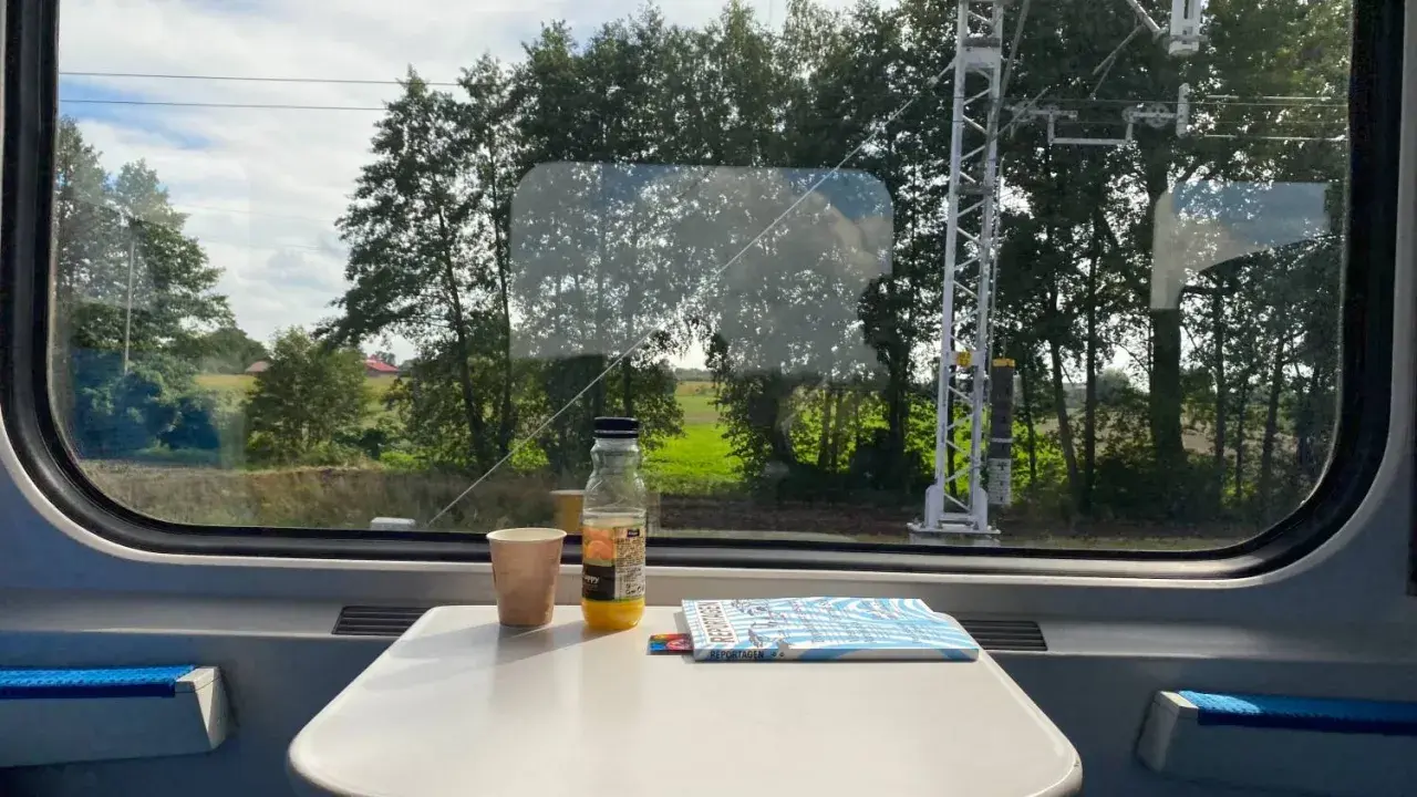 Blick aus dem Zugfenster auf die vorbeiziehende Landschaft. Auf dem Tisch stehen ein Becher, eine Saftflasche und ein Magazin. Die Reise von Warschau nach Berlin beginnt.