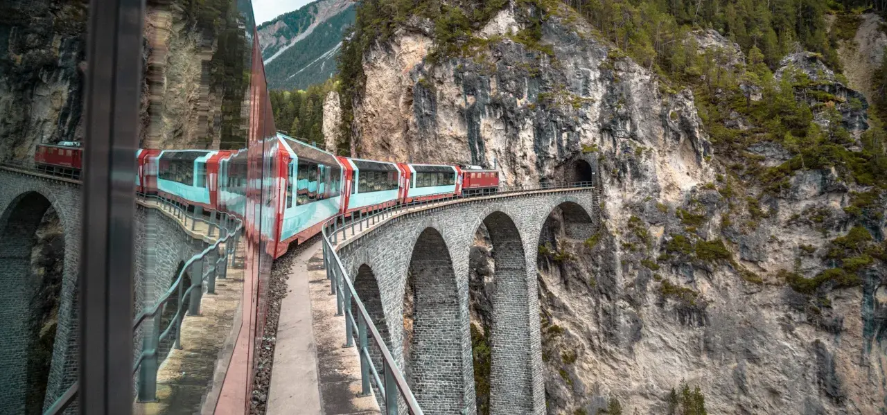 Roter Zug fährt über eine Steinbogenbrücke in den Schweizer Alpen. Perfekt für eine Schweiz Rundreise Zug.
