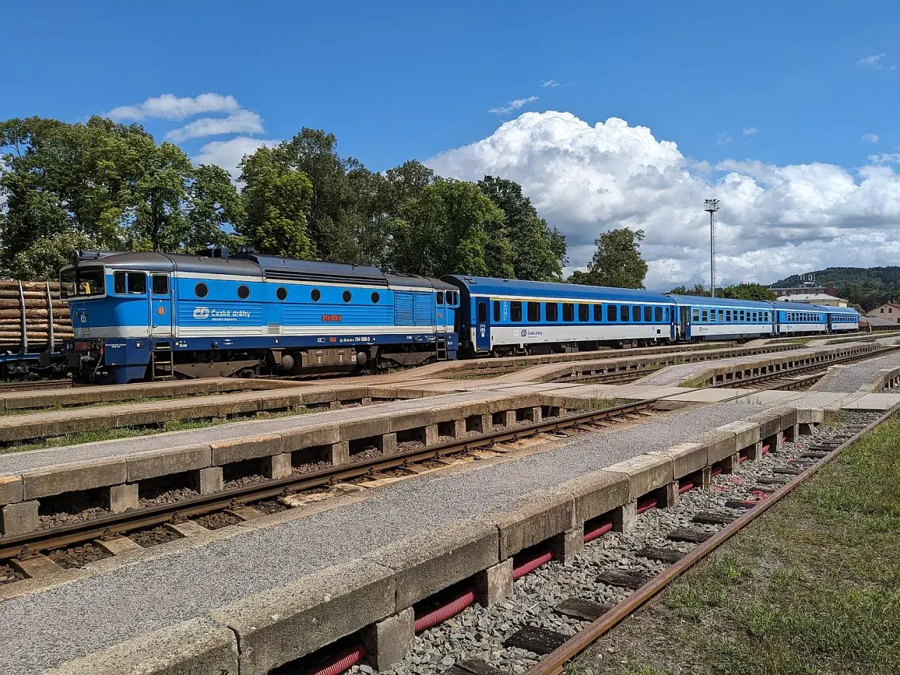 Blauer cz train mit mehreren Waggons steht auf Gleisen, umgeben von Bäumen und einem blauen Himmel mit Wolken.
