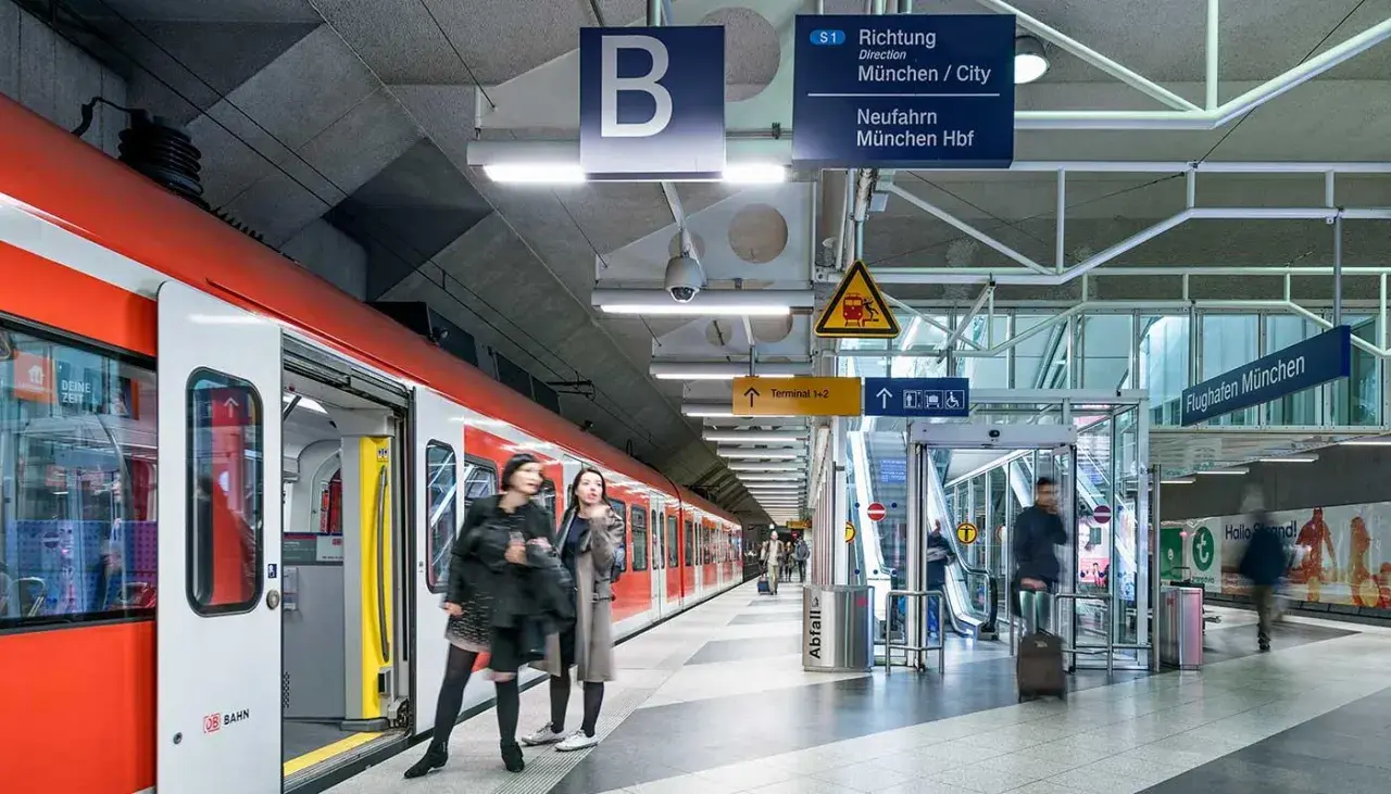 Rote S-Bahn am Bahnsteig, bereit für die Fahrt zum Flughafen München. Schilder weisen den Weg.