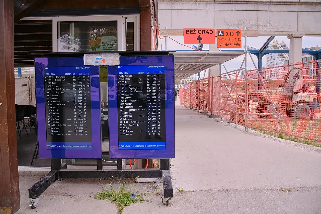 Anzeigetafeln am Bahnhof Ljubljana zeigen Abfahrts- und Ankunftszeiten. Baustellenabsperrungen und ein Bagger sind im Hintergrund sichtbar.