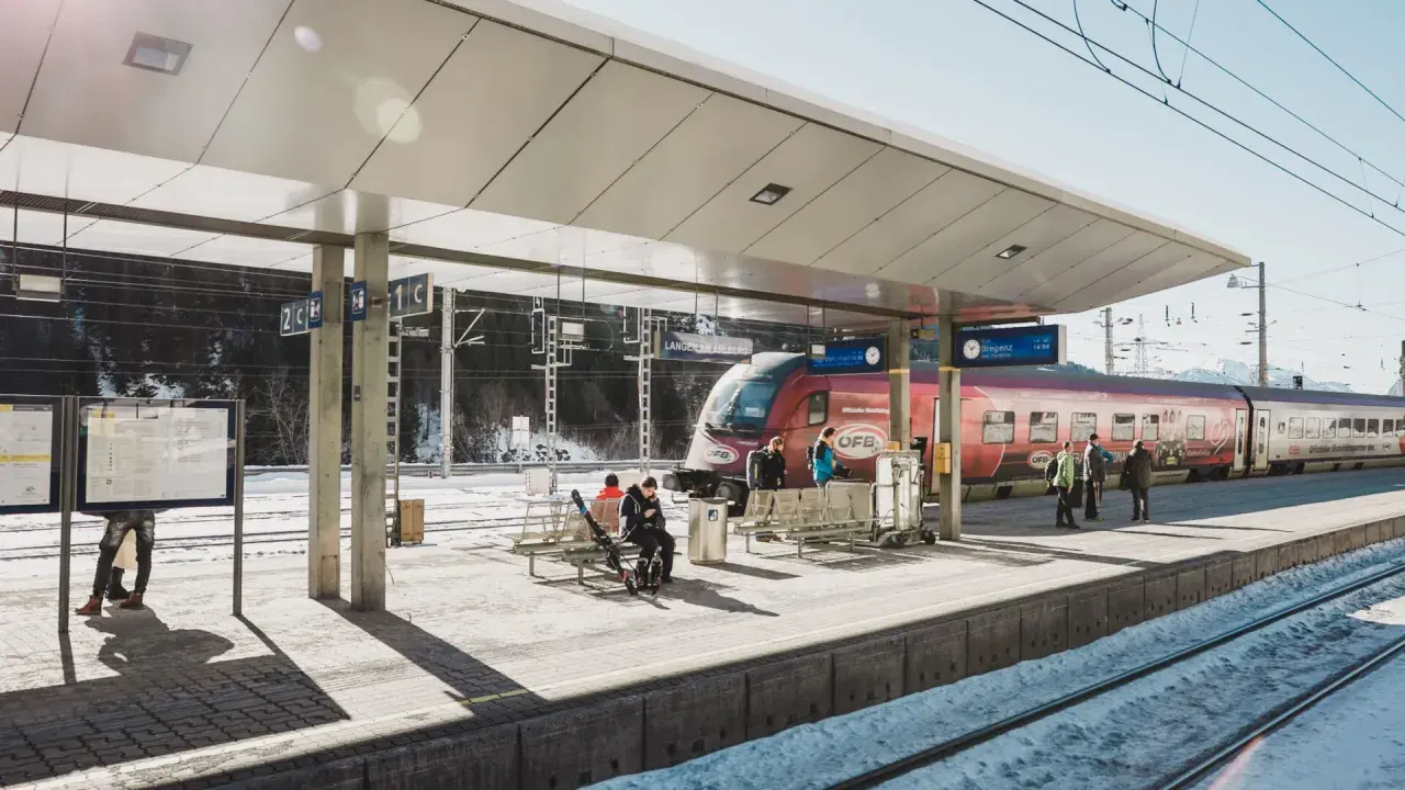Winterliche Szene am **Lech Bahnhof**. Ein roter Zug steht bereit, während Reisende auf dem verschneiten Bahnsteig warten.