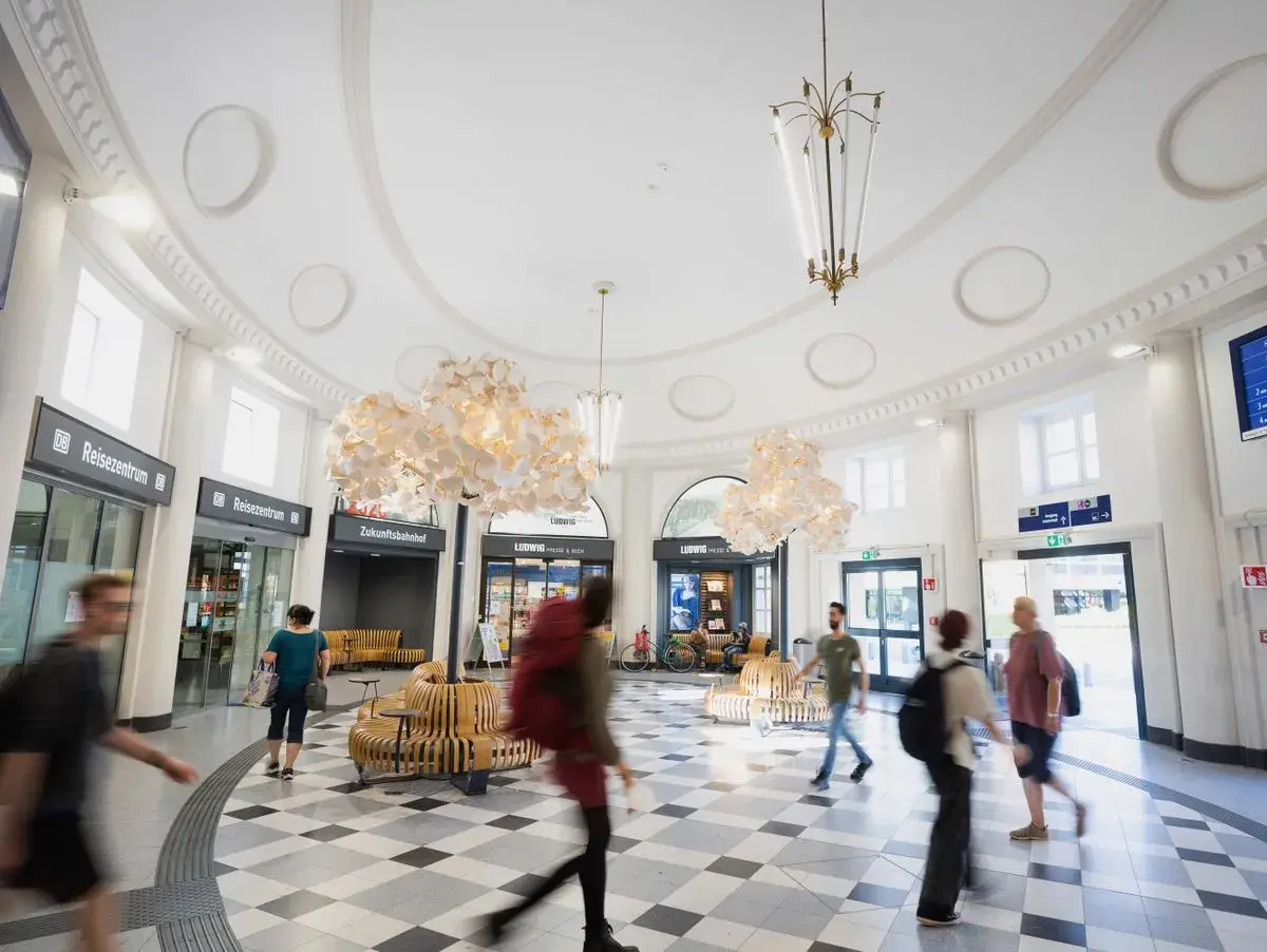 Menschen eilen durch den hellen, modernen **Coburger Bahnhof** mit seinen auffälligen Lampen und Sitzgelegenheiten.