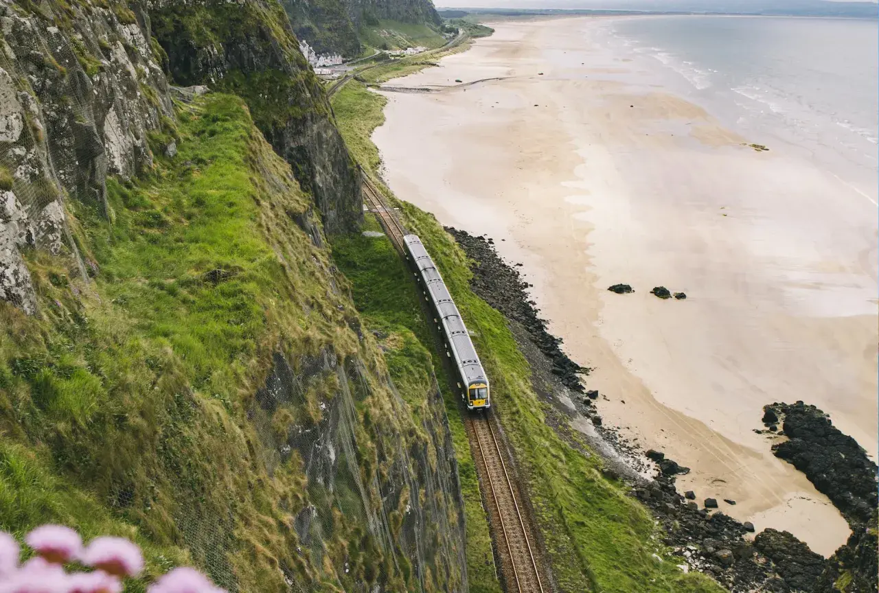 Ein Zug fährt entlang der Küste von **Railway Ireland**. Grüne Hänge stürzen zum Strand ab, das Meer glitzert in der Ferne.