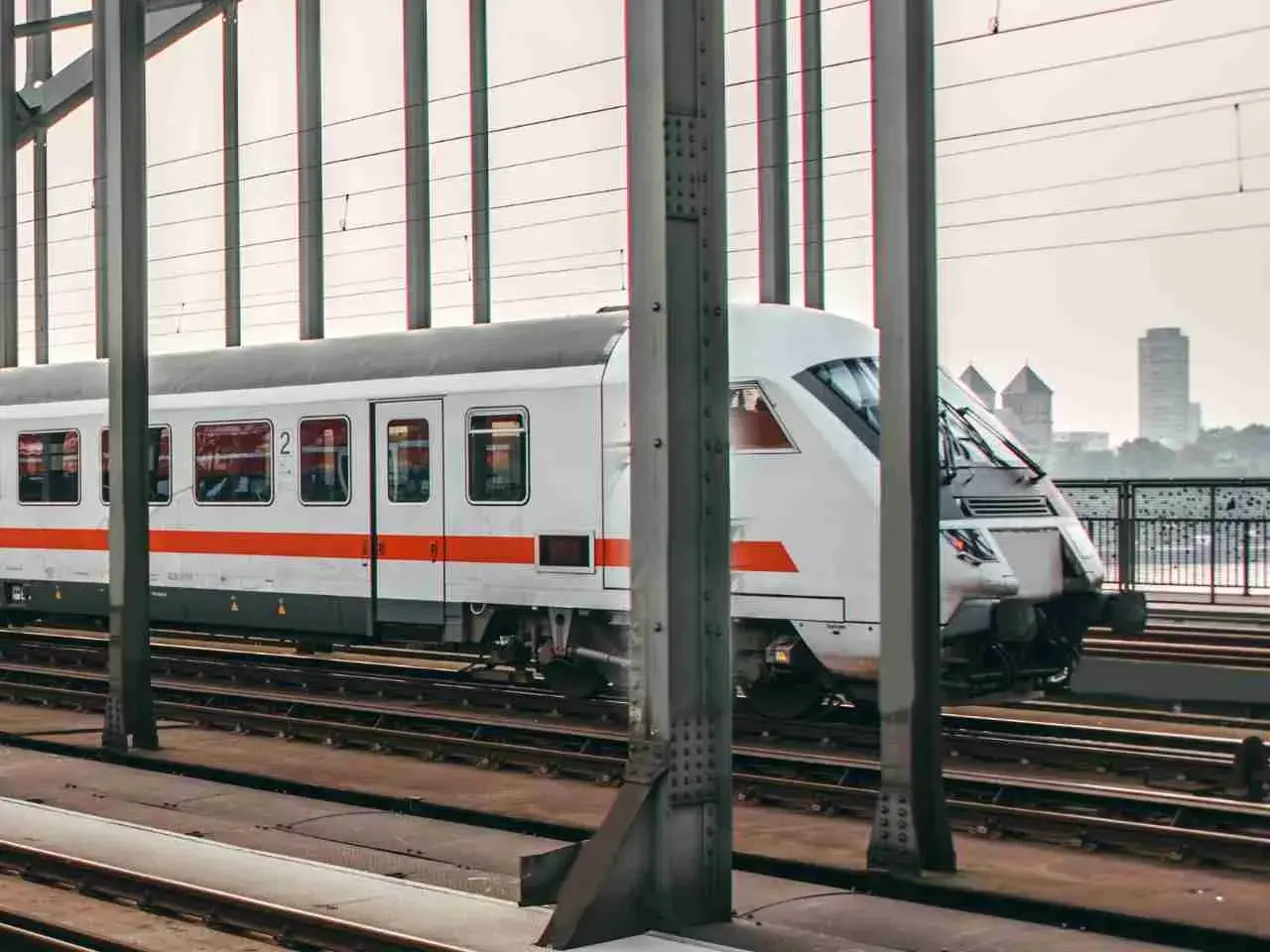 Moderner weißer IC-Zug mit orangefarbenen Streifen fährt auf Gleisen über eine Brücke. Im Hintergrund ist eine Stadt zu sehen.