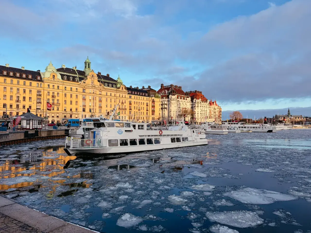 Eisbedeckter Kanal in Stockholm mit einem weißen Schiff, das an einer Promenade mit historischen Gebäuden vorbeifährt. Ein blauer Bus, der an einen schwedischen Zug erinnert, ist im Hintergrund zu sehen.