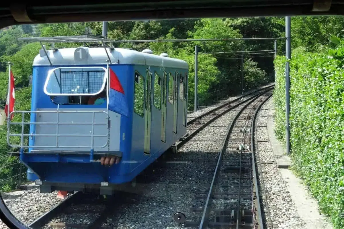 Mit dem Zug zum Lago Maggiore: Eine blaue Standseilbahn fährt auf Schienen durch üppiges Grün.