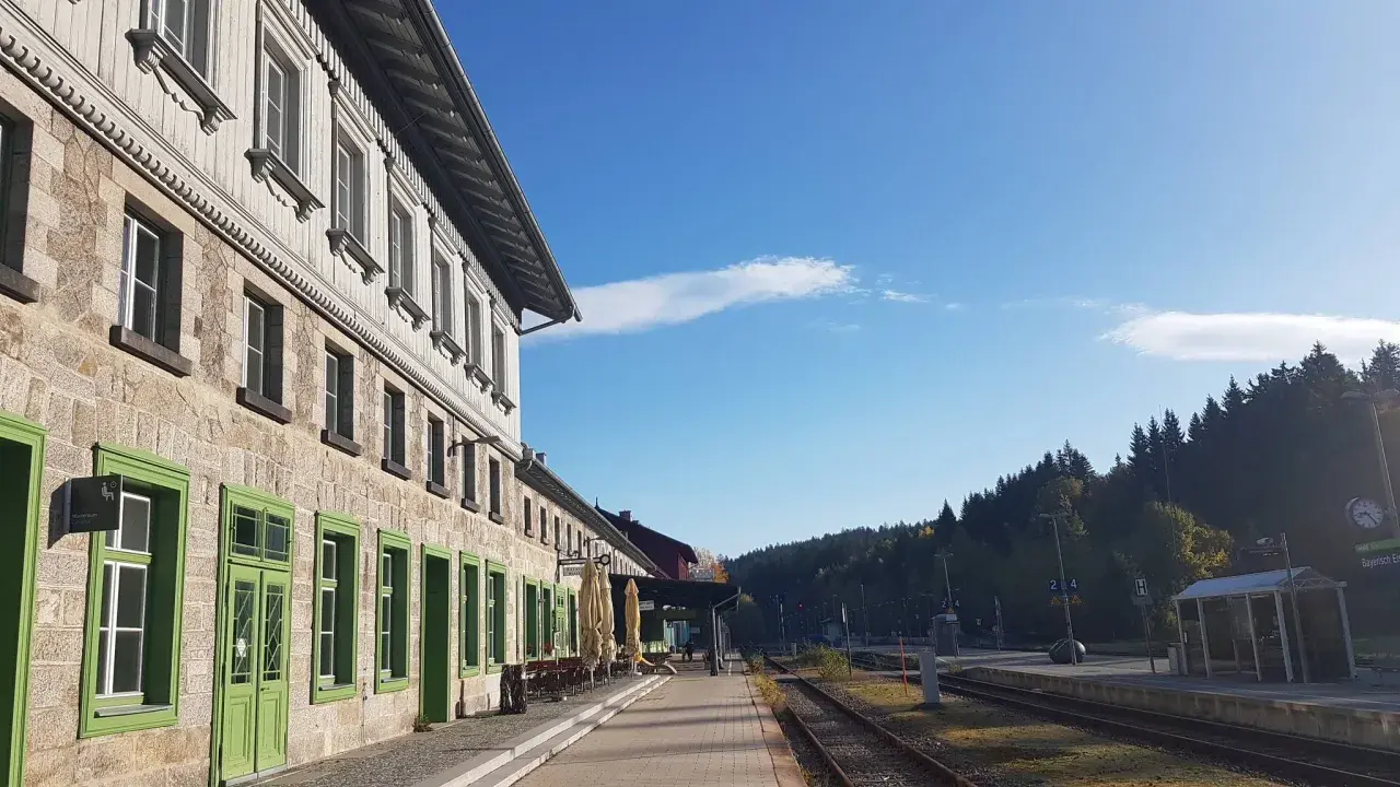 Der **Bayerisch Eisenstein Bahnhof** mit seinem historischen Gebäude und den grünen Fensterrahmen, umgeben von einer Waldkulisse unter blauem Himmel.