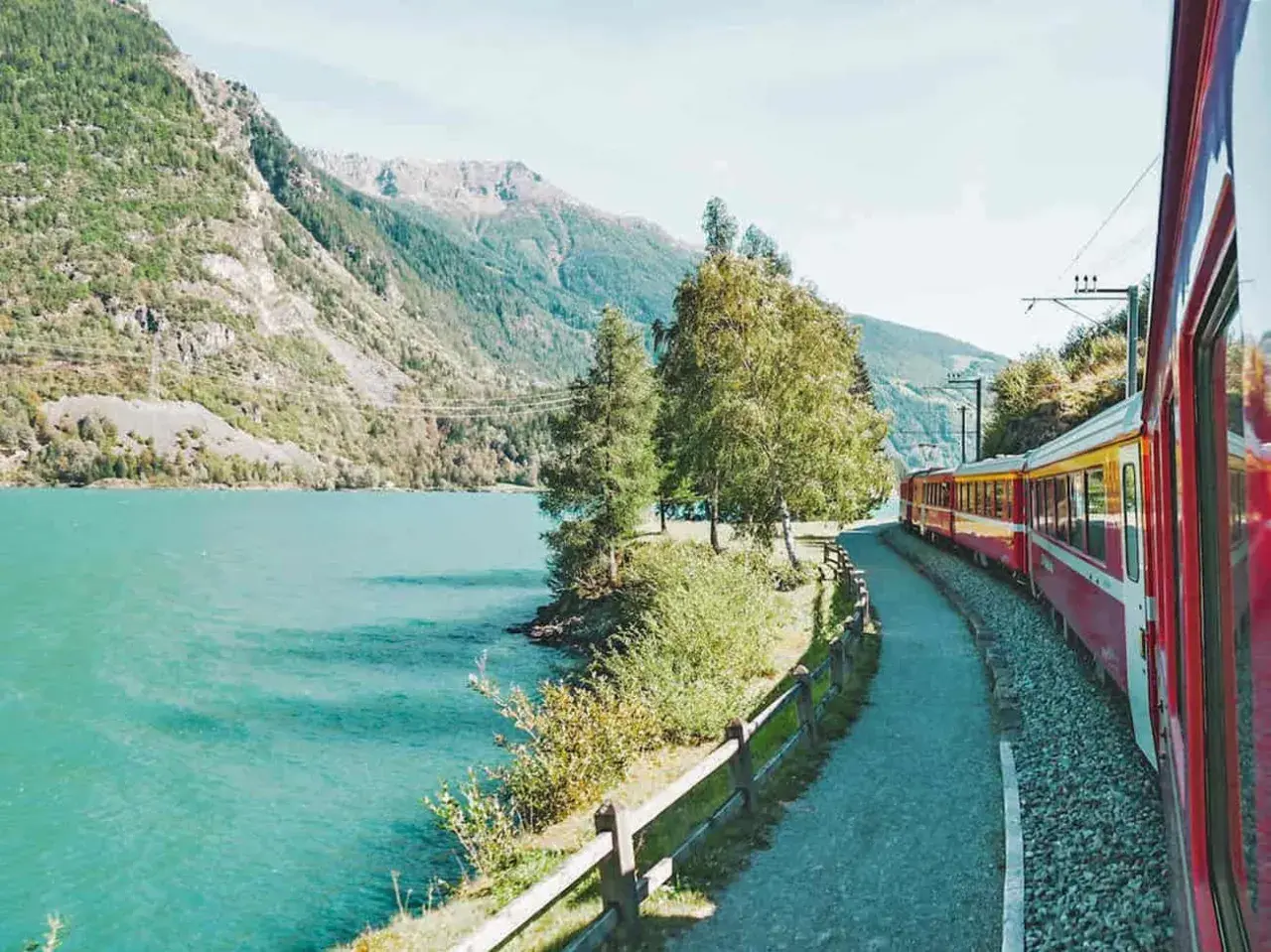Ein roter und gelber Fernzug fährt entlang eines türkisfarbenen Sees und majestätischer Berge.