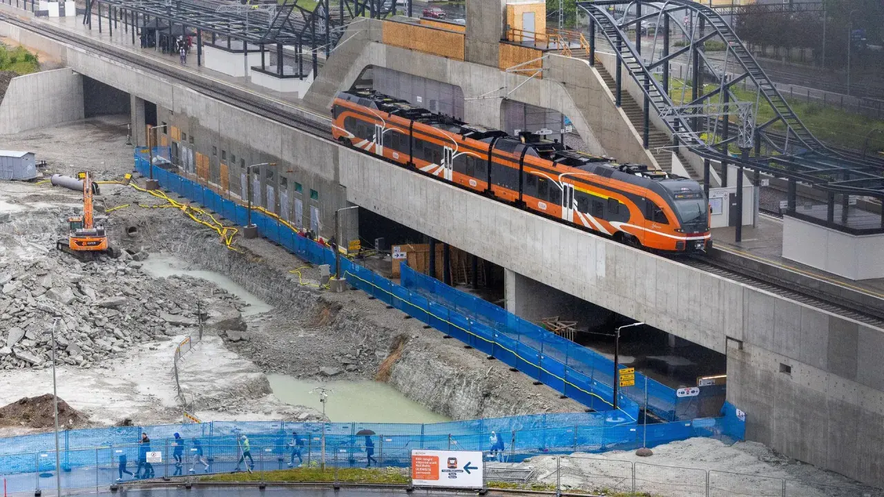 Moderner orange-schwarzer estonia train fährt auf einer erhöhten Strecke, während unten Bauarbeiten mit Baggern und blauen Absperrungen stattfinden.