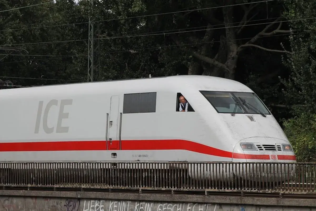 Ein Mann im Führerhaus eines weißen ICE-Zuges mit rotem Streifen. Der ICE-Service fährt auf einer Brücke.