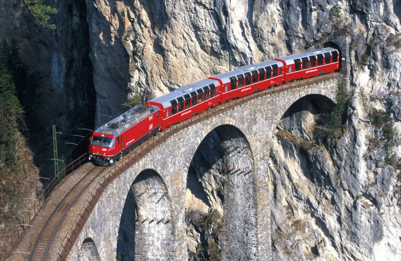 Roter Zug fährt über eine Steinbrücke in den Alpen, auf dem Weg von München nach Italien.