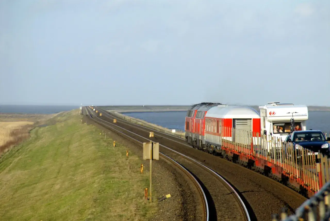 Ein roter Zug f&auml;hrt auf einem Bahndamm mit Blick auf das Meer. Autos und ein Wohnmobil sind auf einem Waggon geladen. Eine Reise mit dem Zug durch Deutschland.