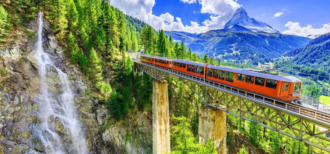 Orange train crosses a viaduct near a waterfall, with the Matterhorn in the background. A scenic journey from Paris to Zurich.