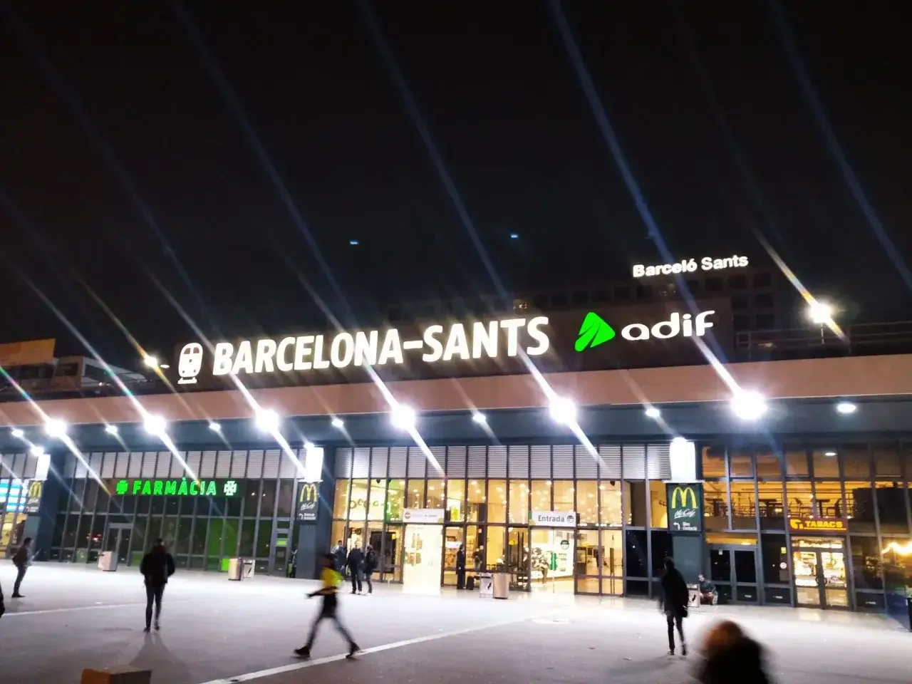 Nachts leuchtet die Barcelona-Sants Hauptbahnhof-Fassade mit den Logos von McDonald's und Farmacia.