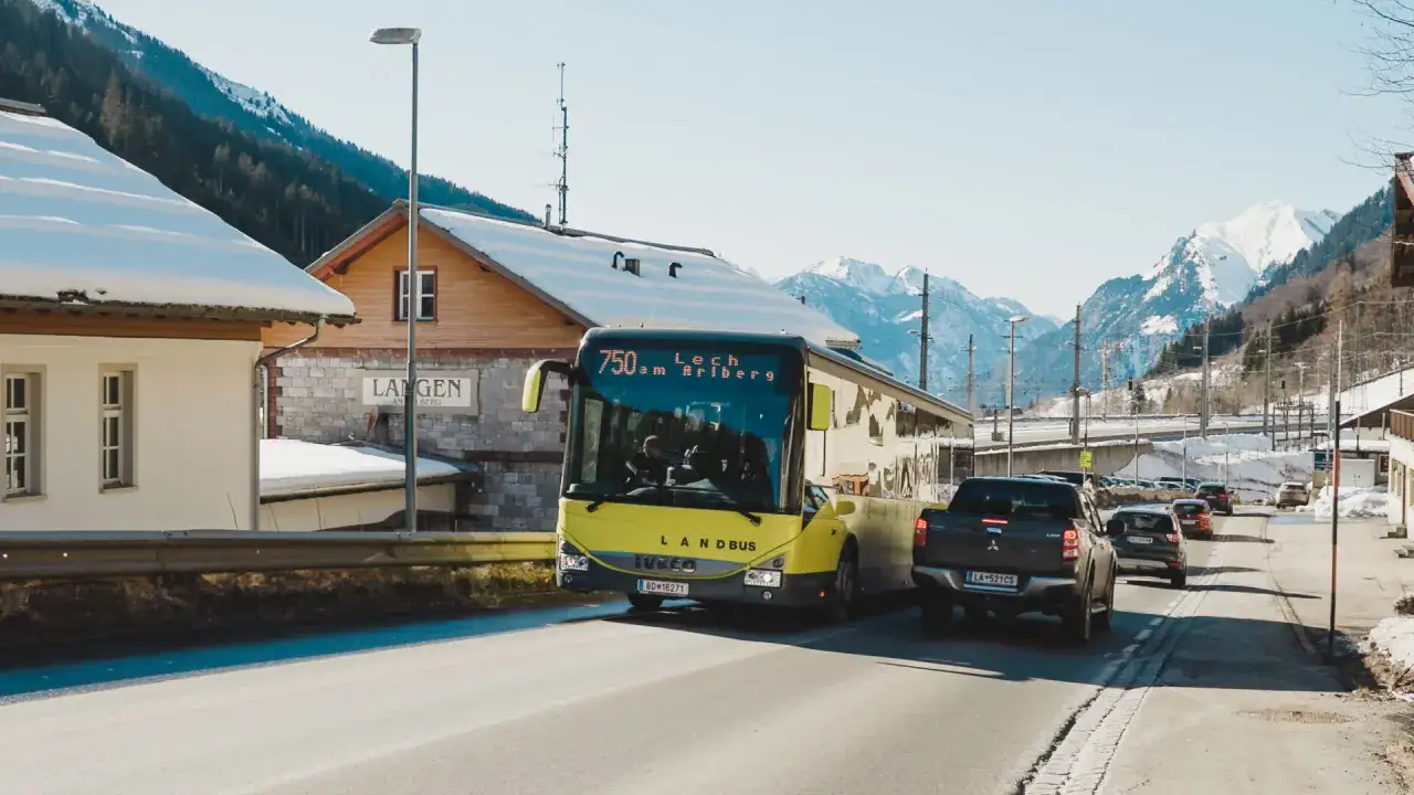Gelber Landbus fährt Richtung Lech am Arlberg. Schneebedeckte Berge im Hintergrund, Langen am Berg im Vordergrund.