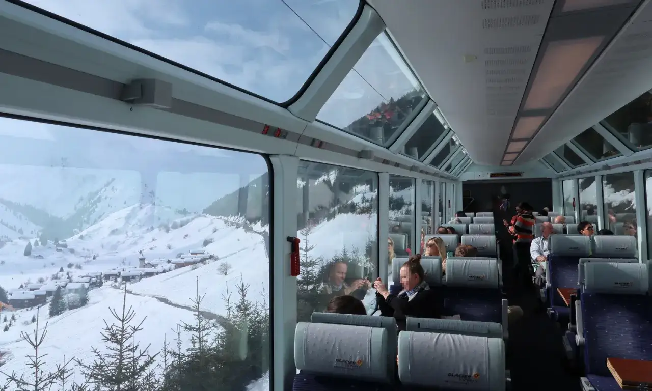 Blick aus dem Panoramawagen eines Glacier Express. Eine Schweiz Rundreise Zug durch verschneite Alpenlandschaft mit kleinen Dörfern.
