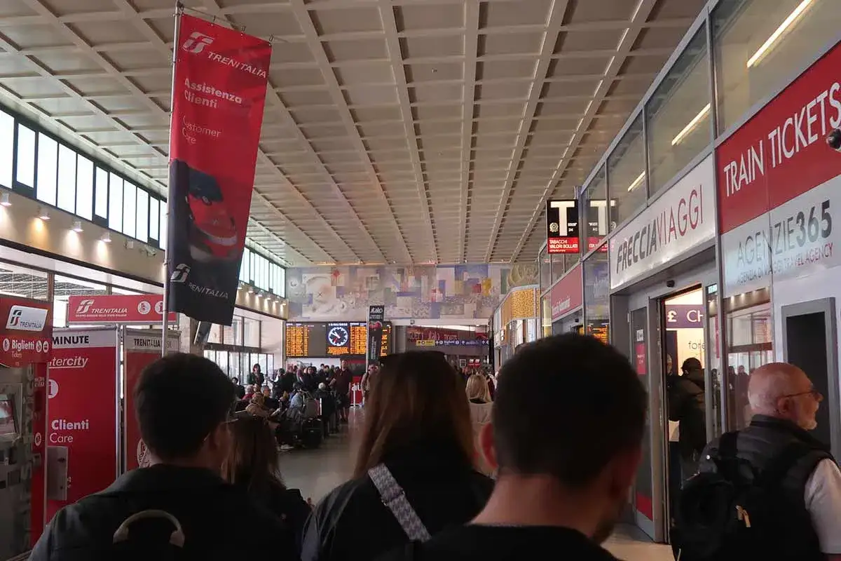 Menschen im Hauptbahnhof Venedig, mit roten Trenitalia-Bannern und Anzeigen für Zugtickets.