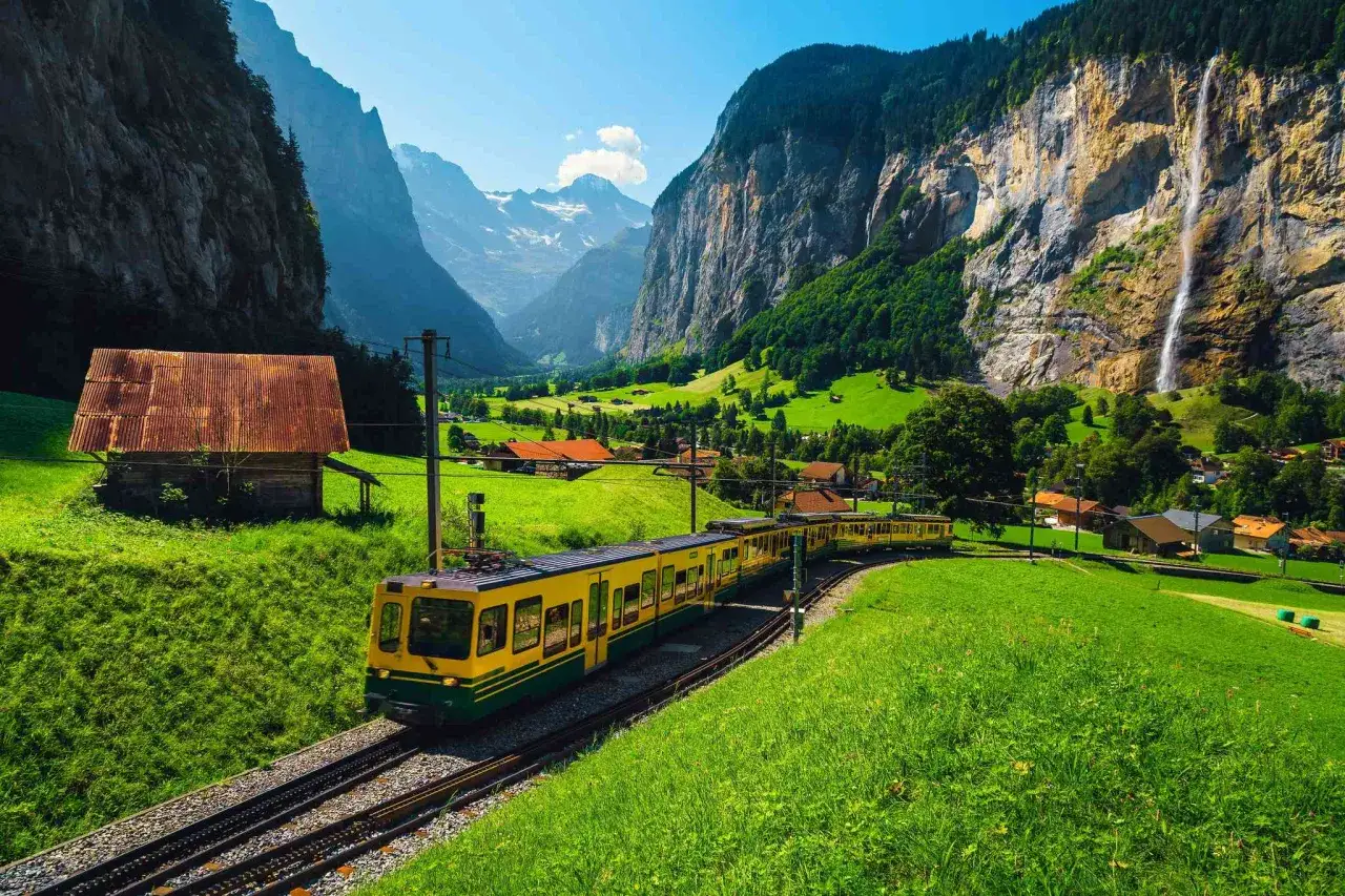 Gelbe Bahn fährt durch grüne Wiesen mit Wasserfall und Bergen, die Reise von Interlaken nach Lauterbrunnen beginnt.