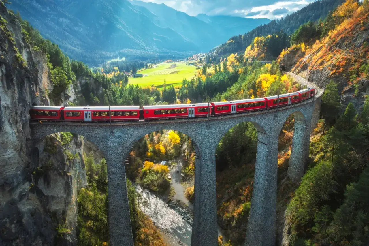 Roter Zug &uuml;berquert eine Steinbr&uuml;cke in den Alpen. Die Herbstfarben leuchten, w&auml;hrend die Fahrt nach Rom beginnt.