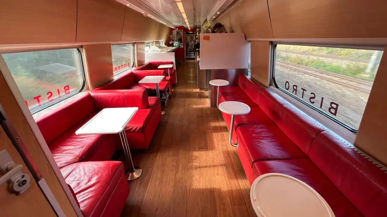 Innenraum eines SJ Intercity Bistrowagens mit roten Ledersitzen und Holzboden. Draußen vorbeiziehende Landschaft.