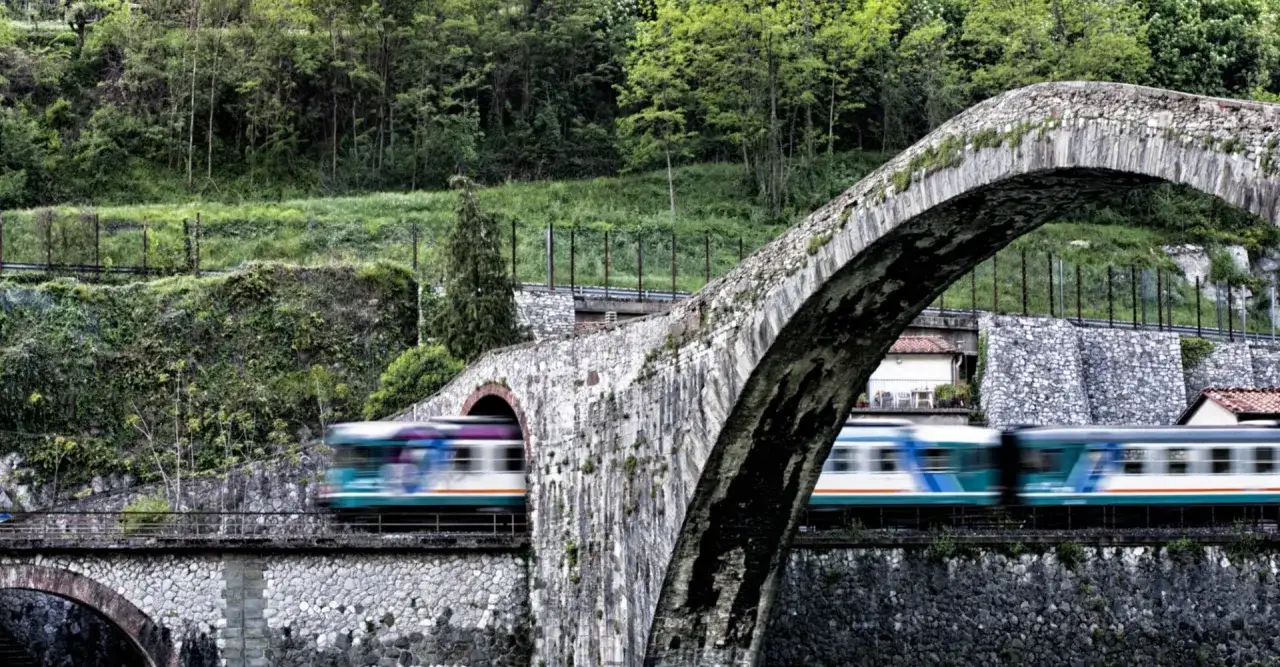 Mit dem Zug in die Toskana: Ein Zug f&auml;hrt &uuml;ber eine alte Steinbr&uuml;cke, umgeben von &uuml;ppigem Gr&uuml;n.