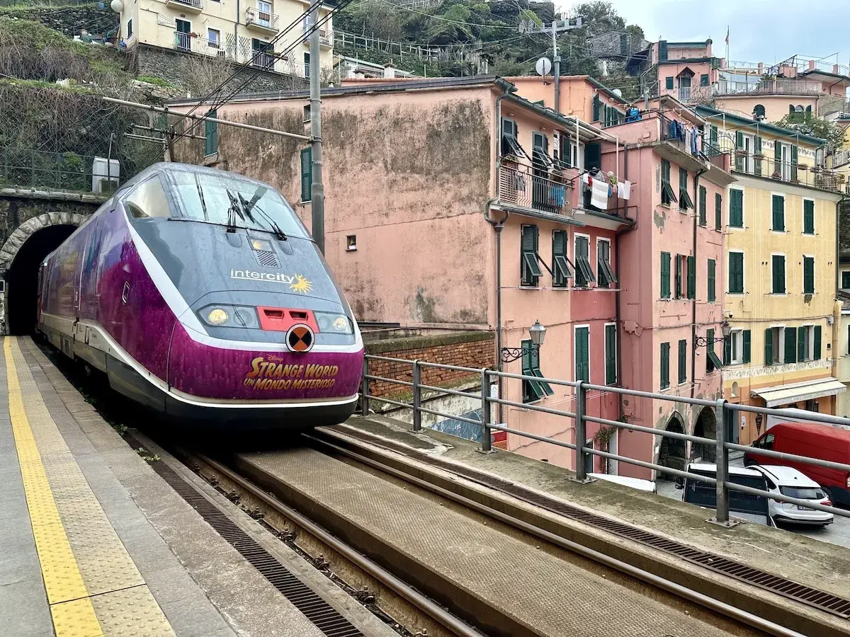 Mit dem Zug durch Italien: Ein bunter Intercity-Zug fährt aus einem Tunnel in eine Küstenstadt mit pastellfarbenen Häusern.